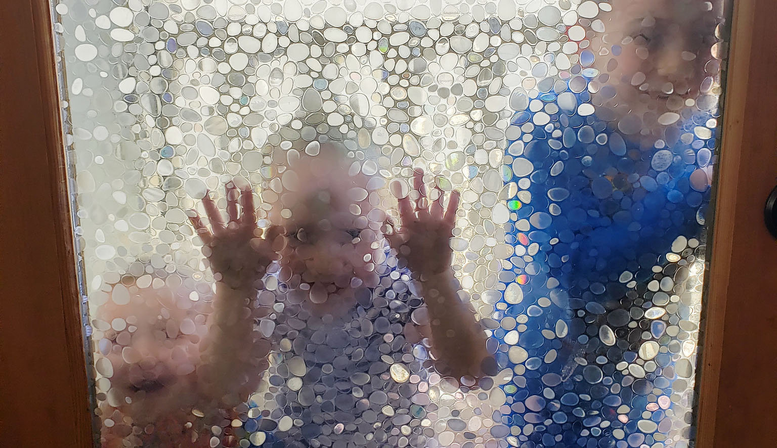 Three kids standing on the outside of a glass door, leaning on the door and pressing their faces and hands to it.