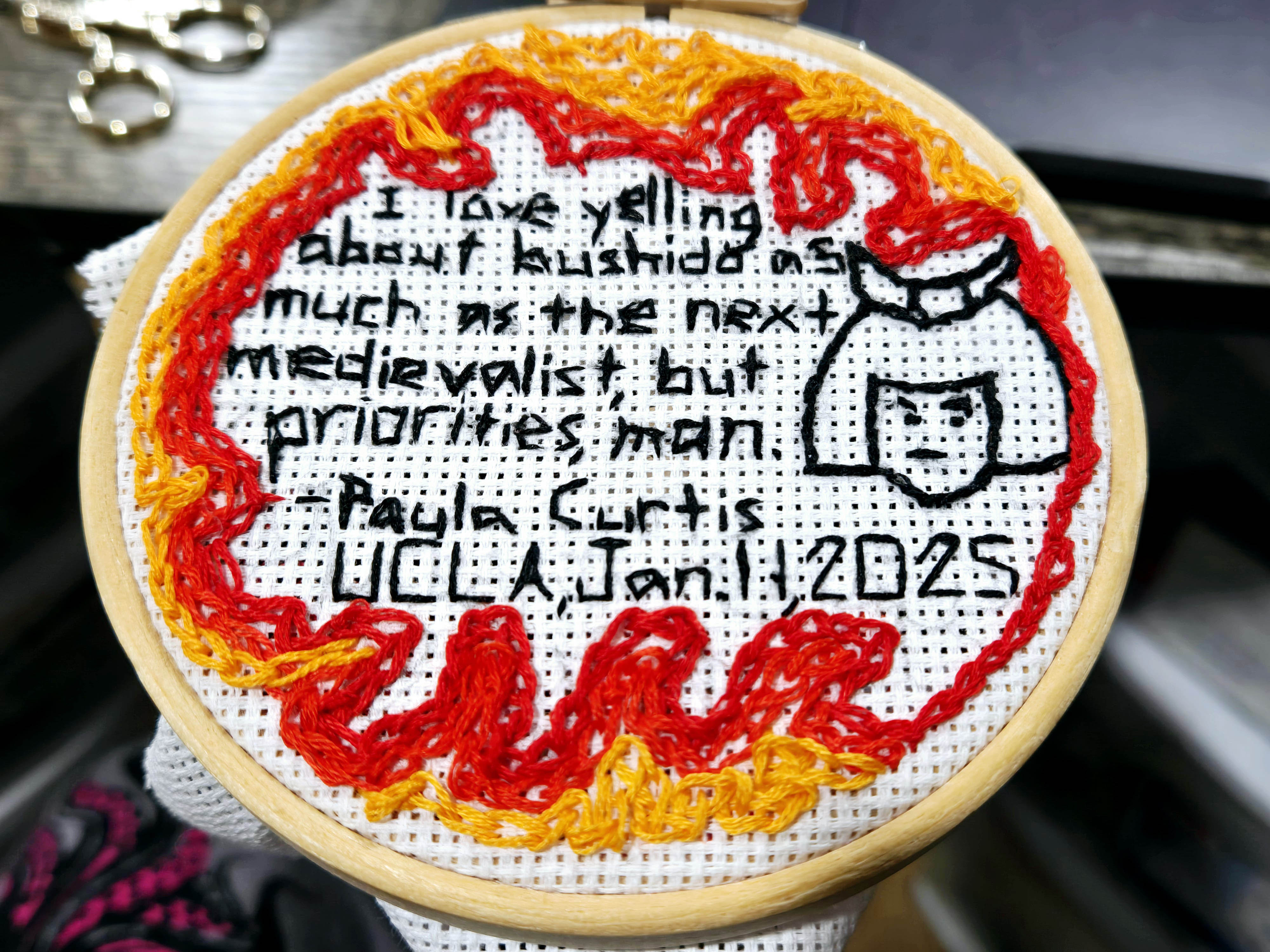 Hand embroidery that reads in black 'I love yelling about bushido as much as the next medievalist, but priorities, man. - Paula Curtis, UCLA, Jan 11, 2025'. There's the head of a samurai embroidered and chain stitch on red, orange, and yellow as flames.