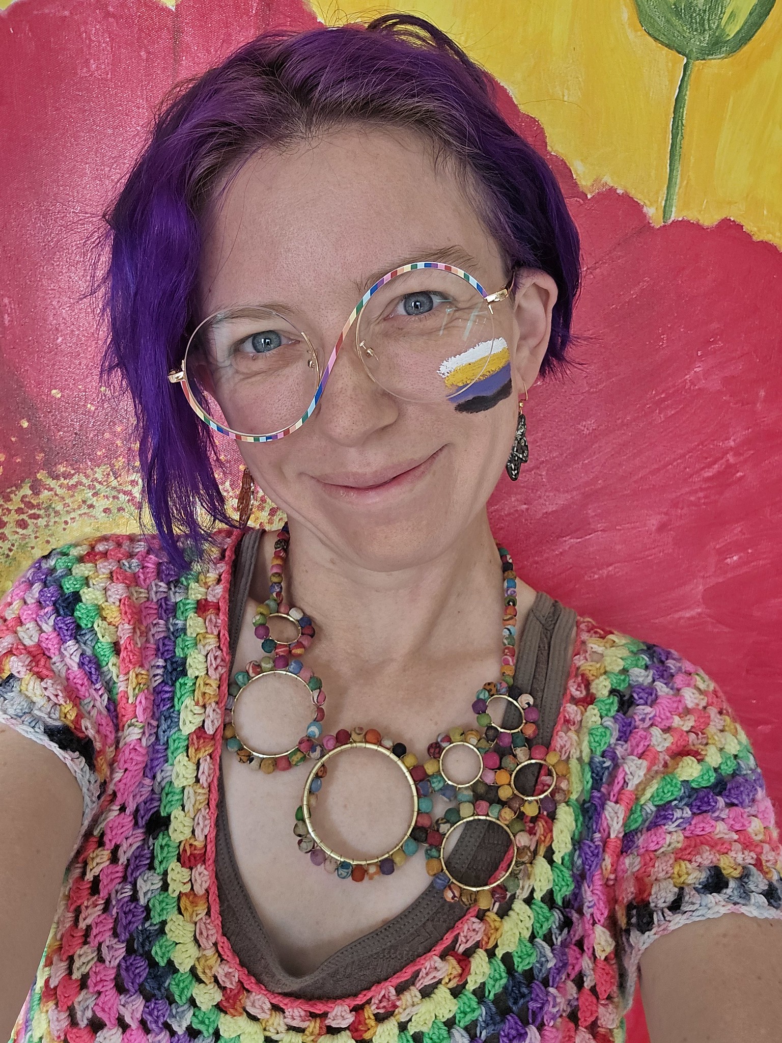 Quinn wearing a rainbow crochet top, rainbow swirl glasses, a rainbow necklace of circles, and a nonbinary pride flag on their cheek.